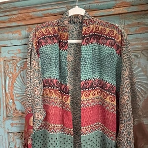 May include: A vibrant, open-front kimono jacket with a patchwork design. The jacket features stripes of teal, red, and patterned fabric with tassels at the hem. The jacket is hanging on a white hanger.