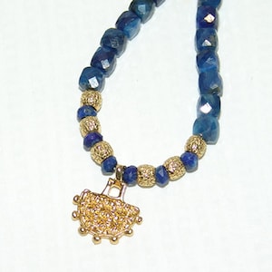 以下が含まれることがあります： 濃い青色のファセットビーズと金色のテクスチャードビーズをあしらったネックレス。 金色のペンダントには花の模様が施されており、ネックレスから吊り下げられています。 ネックレスは白い背景に表示されています。