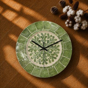 Puede incluir: Un reloj de pared redondo con un borde de mosaico de azulejos verdes y una esfera central de color crema decorada con un patrón radial de azulejos verdes en forma de hoja. El reloj tiene tres manecillas negras finas en el centro.