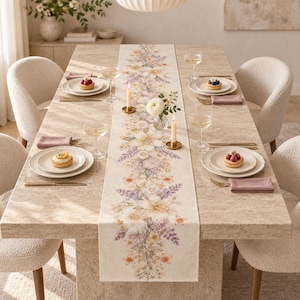 Può includere: Un runner da tavola rettangolare color beige con una stampa botanica centrale di vari fiori nelle tonalità del viola, arancione e crema. Il runner è posizionato su un tavolo da pranzo in pietra con quattro piatti bianchi, ognuno con una crostatina.
