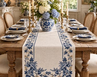 Könnte beinhalten: Ein beigefarbener Tischläufer aus Leinen mit blau-weißem Blumenmuster und Pagodenmotiven, mittig auf einem hölzernen Esstisch platziert. Der Läufer ist mit detaillierten botanischen Mustern verziert und von Gedecken mit blauen Servietten umgeben.