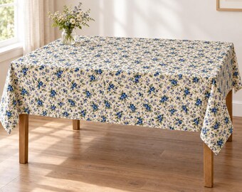 Könnte beinhalten: Eine rechteckige Tischdecke mit cremefarbenem Untergrund und einem dichten, sich wiederholenden Muster aus kleinen blauen Blumen und grünen Blättern. Eine klare Glasvase mit einem Strauß weißer und blauer Wildblumen steht auf dem Tisch.