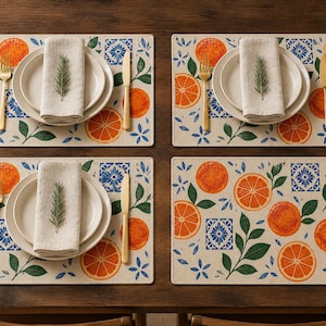 May include: Four rectangular placemats featuring a pattern of orange slices, green leaves, and blue geometric tiles on a cream background. Each setting includes a white plate, a folded beige napkin topped with a rosemary sprig, a gold fork, and a gold knife.