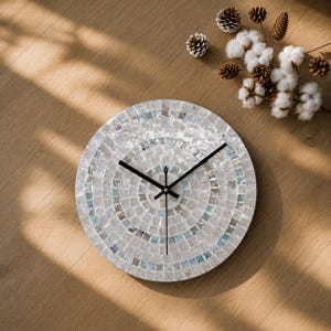 Peut inclure: Une horloge murale circulaire dotée d'un cadran en mosaïque composé de carreaux irisés et nacrés dans des tons de blanc, de crème et de bleu doux. L'horloge possède trois fines aiguilles en métal noir placées au centre pour indiquer l'heure.