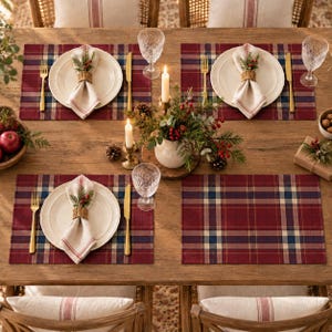 Peut inclure: Une table à manger en bois dressée pour quatre personnes avec des sets de table à carreaux rouges et bleus, des assiettes en céramique crème, des serviettes en lin avec des ronds de serviette tressés, des couverts dorés et des verres.