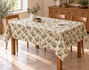 Könnte beinhalten: Eine rechteckige Tischdecke mit cremefarbenem Hintergrund und einem sich wiederholenden Muster aus grünen Olivenzweigen, dunkelblauen Oliven und kleinen weißen Blüten, die über einem hölzernen Esstisch mit vier passenden Holzstühlen liegt.