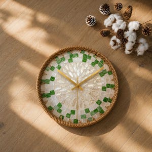 Puede incluir: Un reloj de pared redondo con una esfera de mosaico hecha de piezas de concha blanca iridiscente y pequeños azulejos cuadrados verdes. El reloj está enmarcado por un borde grueso de cuerda de yute natural trenzada y manecillas de metal dorado.