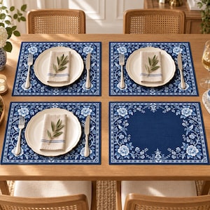 Può includere: Quattro tovagliette rettangolari con sfondo blu scuro e un bordo con motivo floreale e di uccelli bianchi. Tre tovagliette sono apparecchiate con un piatto in ceramica bianca, un tovagliolo beige piegato con strisce blu, una forchetta e un coltello d'argento.