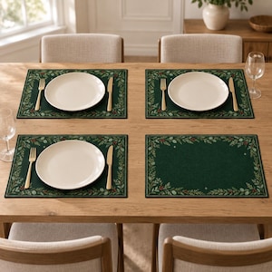 May include: A set of four rectangular dark green placemats featuring a decorative border of pine branches, holly berries, and small stars. Three of the placemats are set with a white ceramic plate, a gold fork, and a gold knife for dining.