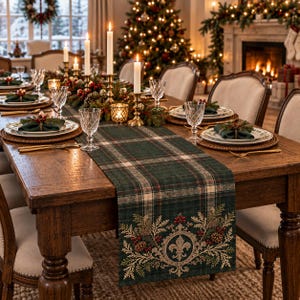 May include: A dark green plaid table runner featuring a central fleur-de-lis emblem surrounded by embroidered pine branches, red berries, and pinecones. The runner is laid across a wooden dining table set with white plates, napkins, and candles.
