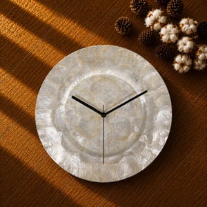 Puede incluir: Un reloj de pared circular con una esfera hecha de discos de concha blancos iridiscentes superpuestos. El reloj tiene tres manecillas negras delgadas posicionadas en el centro. La superficie de la esfera muestra una textura natural nacarada con varios tonos de blanco roto y crema, creando una apariencia reflectante y estratificada.