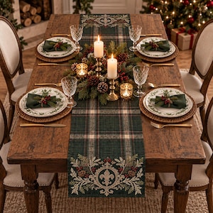 May include: A dark green plaid table runner featuring a central fleur-de-lis motif, pine branches, and red berries. The runner is displayed on a wooden dining table set with white plates, green napkins, gold cutlery, and two lit pillar candles.