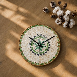 Puede incluir: Un reloj de pared redondo con una esfera de mosaico hecha de piezas de piedra blancas y verdes. El centro muestra un patrón floral verde rodeado por un anillo de triángulos oscuros. El reloj tiene manecillas negras y un borde trenzado.