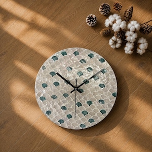 May include: A round wall clock featuring a mosaic pattern of iridescent white mother-of-pearl and teal-colored shell pieces arranged in a scalloped design. The clock has three thin black hands centered on the face with no visible numbers or markers.