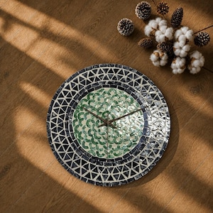 May include: A round wall clock featuring a mosaic border of dark gray and black triangular tiles. The center displays a circular pattern of iridescent green, crescent-shaped tiles. Two thin, dark metal clock hands are positioned in the center.