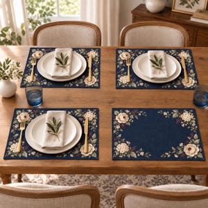 Può includere: Un set di quattro tovagliette rettangolari blu navy con un bordo floreale di peonie color crema e foglie verdi. Tre coperti includono un piatto bianco, un tovagliolo beige piegato con un rametto d'ulivo, una forchetta e un coltello dorati.