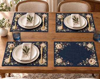 Könnte beinhalten: Ein Set aus vier rechteckigen, marineblauen Platzsets mit einem Blumenrand aus cremefarbenen Pfingstrosen und grünen Blättern. Drei Gedecke umfassen einen weißen Teller, eine gefaltete beige Serviette mit Olivenzweig, eine goldene Gabel und ein goldenes Messer.