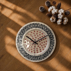 Può includere: Un orologio da parete rotondo con un quadrante centrale color crema decorato con piccoli motivi a fiori rosa. L'orologio è incorniciato da un ampio bordo di tessere di mosaico triangolari grigio scuro e bianche e ha tre sottili lancette nere.