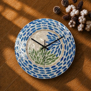 May include: A round wall clock featuring a mosaic design made of small, petal-shaped tiles. The outer border consists of blue tiles, while the centre displays a pattern of green and white tiles. Three thin black hands are centred on the clock face.