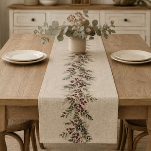 May include: A rectangular beige table runner featuring a central printed design of green pine branches and red berries. The runner is laid flat across a wooden dining table, centered between two white ceramic plates and a small vase of greenery.
