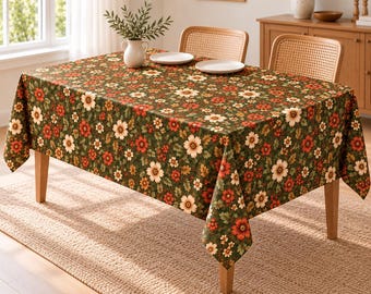 Könnte beinhalten: Eine rechteckige Tischdecke mit einem dichten Blumenmuster aus cremefarbenen, orangefarbenen und roten Blüten auf dunkelgrünem Grund mit grünen Blättern. Die Tischdecke liegt auf einem Holztisch mit zwei hellen Stühlen.