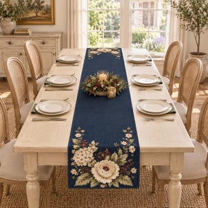 Puede incluir: Un camino de mesa rectangular de color azul marino con un diseño floral estampado que presenta grandes peonías blancas, flores más pequeñas y hojas verdes en ambos extremos, colocado sobre una mesa de comedor de madera clara.