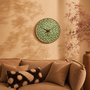 Op de afbeelding: Een ronde wandklok met een groen en wit bladpatroon op de wijzerplaat en dunne zwarte wijzers, gemonteerd aan een beige muur boven een gestructureerde beige bank met een bruin en beige rechthoekig kussen en een beige vierkant kussen.