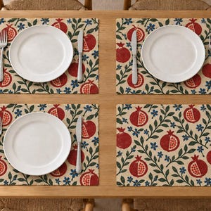 Op de afbeelding: Een set van vier rechthoekige placemats met een herhalend patroon van rode granaatappels, groene bladeren en kleine blauwe bloemen op een beige achtergrond. Elke placemat is gedekt met een wit bord, een zilveren vork en een zilveren mes.