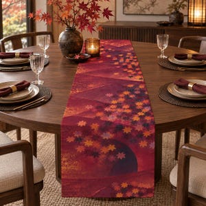 May include: A rectangular table runner featuring a deep red and burgundy gradient background decorated with a pattern of scattered autumn maple leaves in shades of orange, yellow, and dark purple, laid across a round wooden dining table.