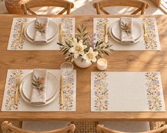Könnte beinhalten: Ein Set aus vier rechteckigen beigen Platzsets mit einem Blumenmuster aus weißen Lilien und kleinen violetten Blüten an den Seiten. Jedes Platzset ist mit einem weißen Keramikteller, einer beigen Leinenserviette, einer goldenen Gabel und einem goldenen Messer gedeckt.