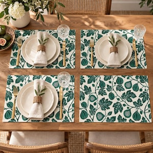 Peut inclure: Un service de table composé de quatre sets de table rectangulaires à motif de feuilles et de fruits de figuier vert foncé sur fond blanc. Chaque couvert comprend une assiette en céramique blanche, une serviette en lin beige et des couverts dorés.