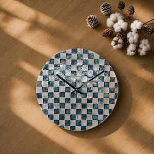 May include: A round wall clock featuring a checkered pattern of alternating blue and iridescent white square tiles. The clock has three thin black hands positioned at the center. The surface of the tiles appears glossy and reflective.