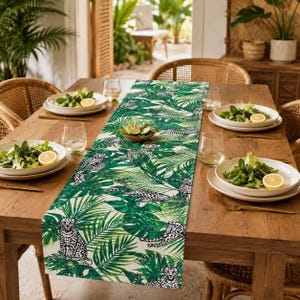 Peut inclure: Un chemin de table rectangulaire orné d'un motif répétitif de léopards et de feuilles de palmier tropicales vert vif sur un fond blanc. Le chemin est centré sur une table à manger en bois avec des assiettes blanches, des salades et des verres.