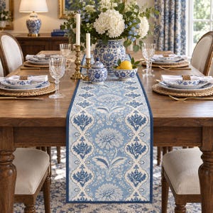 Puede incluir: Un camino de mesa rectangular con un patrón floral de damasco en azul claro y blanco con un borde azul oscuro. El camino está centrado en una mesa de comedor de madera decorada con platos de patrón azul y blanco y copas de cristal.