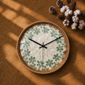 May include: A round wall clock with a woven rattan frame and a cream-coloured face. The clock features a repeating pattern of green flower-shaped motifs around the perimeter and a central starburst design made of white shell-like pieces.