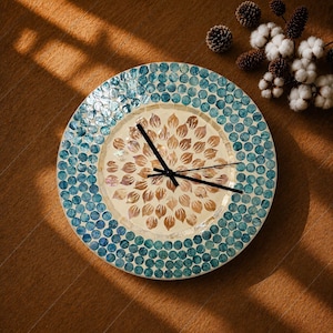 Peut inclure: Une horloge murale ronde avec une bordure en mosaïque composée de pièces circulaires en coquillage bleu irisé. Le cadran central présente un motif radial de pétales en coquillage brun clair sur fond crème avec trois aiguilles noires.
