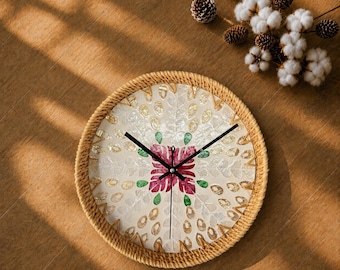 Könnte beinhalten: Eine runde Wanduhr mit einem Rand aus natürlichem Juteseil und einem Zifferblatt, das mit schillernden Perlmuttstücken eingelegt ist. Die Mitte zeigt ein florales Muster in tiefen Rosa- und Grüntönen, ergänzt durch drei schwarze Zeiger.