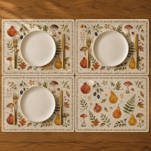 May include: Four rectangular placemats featuring a botanical print of mushrooms, pears, pomegranates, and autumn leaves. Each placemat is set with a plain white ceramic plate, a gold-toned fork on the left, and a gold-toned knife on the right side.