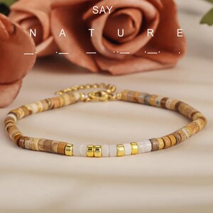 May include: A beaded bracelet featuring small, flat, circular earth-toned stone beads in shades of tan, brown, and beige, interspersed with smooth white beads and polished gold-tone metal spacers. The bracelet includes a gold-tone metal lobster clasp.
