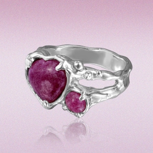 Puede incluir: Un anillo de plata con una piedra preciosa rosa oscuro en forma de corazón y una piedra redonda más pequeña. El anillo tiene un diseño orgánico y texturizado con un acabado pulido. Las piedras están engastadas en un engaste calado detallado.
