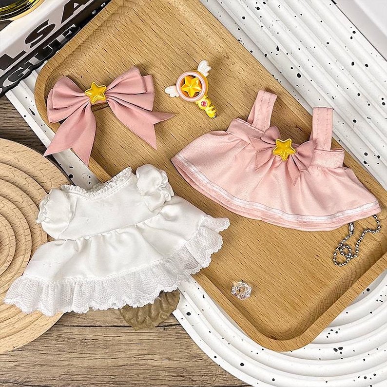 K&ouml;nnte beinhalten: Eine Sammlung von Puppenzubeh&ouml;r, darunter eine rosa Schleife mit gelbem Stern, ein rosa Kleid mit gelbem Stern, ein wei&szlig;es Kleid mit Spitzenbesatz und ein sternf&ouml;rmiger Zauberstab. Die Artikel sind auf einem Holztablett angeordnet.