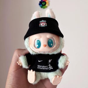 K&ouml;nnte beinhalten: Ein Pl&uuml;schtier-Hase mit schwarzem Visier und schwarzem Trikot mit dem Liverpool-Logo und dem Text "Standard Chartered". Der Hase hat gro&szlig;e blaue Augen und wird in einer Hand gehalten.