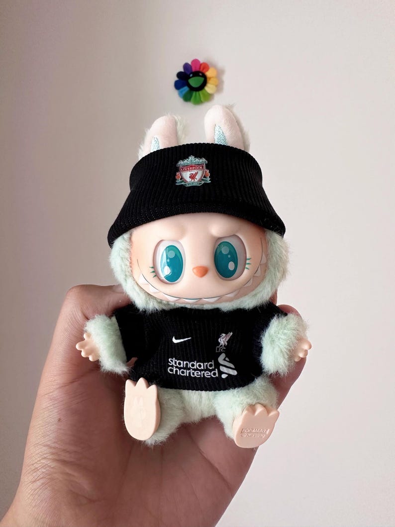 K&ouml;nnte beinhalten: Ein Pl&uuml;schtier-Hase mit schwarzem Visier und schwarzem Trikot mit dem Liverpool-Logo und dem Text "Standard Chartered". Der Hase hat gro&szlig;e blaue Augen und wird in einer Hand gehalten.