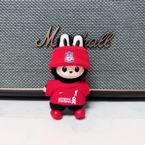 K&ouml;nnte beinhalten: Eine kleine Pl&uuml;schpuppe, die einen roten Liverpool FC-Hut und ein Trikot tr&auml;gt. Die Puppe hat einen schwarzen K&ouml;rper, ein rundes Gesicht und rote Schuhe. Das Trikot zeigt das Nike-Logo und das Standard Chartered-Logo.