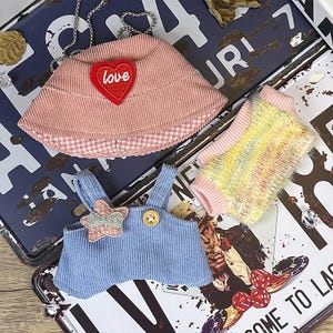 K&ouml;nnte beinhalten: Eine Sammlung von Miniatur-Haustierkleidung. Enth&auml;lt einen rosa Cordhut mit einem "Love"-Herz, eine blaue Cord-Latzhose und einen bunten Pullover. Die Artikel werden auf einem Vintage-Kennzeichen pr&auml;sentiert.