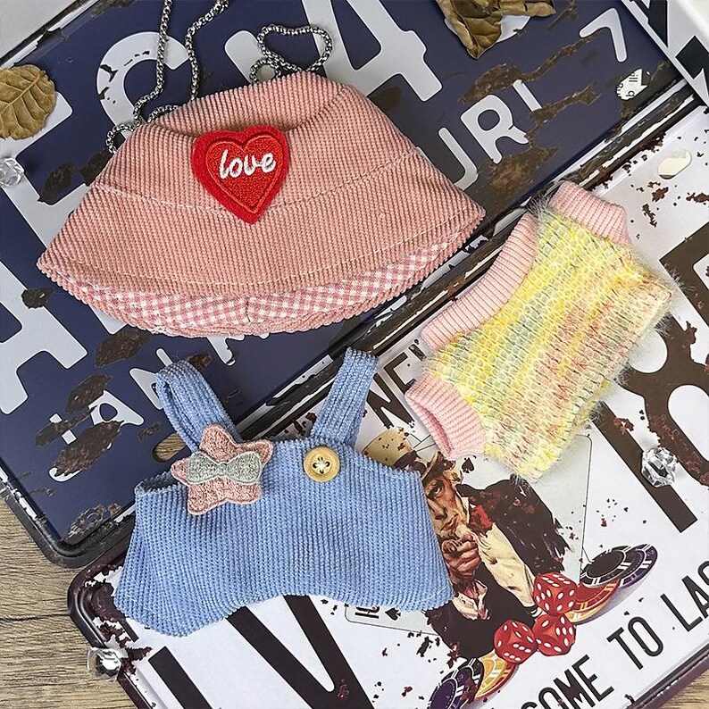 K&ouml;nnte beinhalten: Eine Sammlung von Miniatur-Haustierkleidung. Enth&auml;lt einen rosa Cordhut mit einem "Love"-Herz, eine blaue Cord-Latzhose und einen bunten Pullover. Die Artikel werden auf einem Vintage-Kennzeichen pr&auml;sentiert.