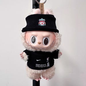 K&ouml;nnte beinhalten: Ein Stofftier-Hase mit schwarzem Strickhut und Pullover mit dem Logo des Liverpool Football Club. Der Pullover zeigt auch das Nike-Logo und den Text "Standard Chartered". Das Spielzeug hat ein hellrosa Gesicht und flauschiges Fell.