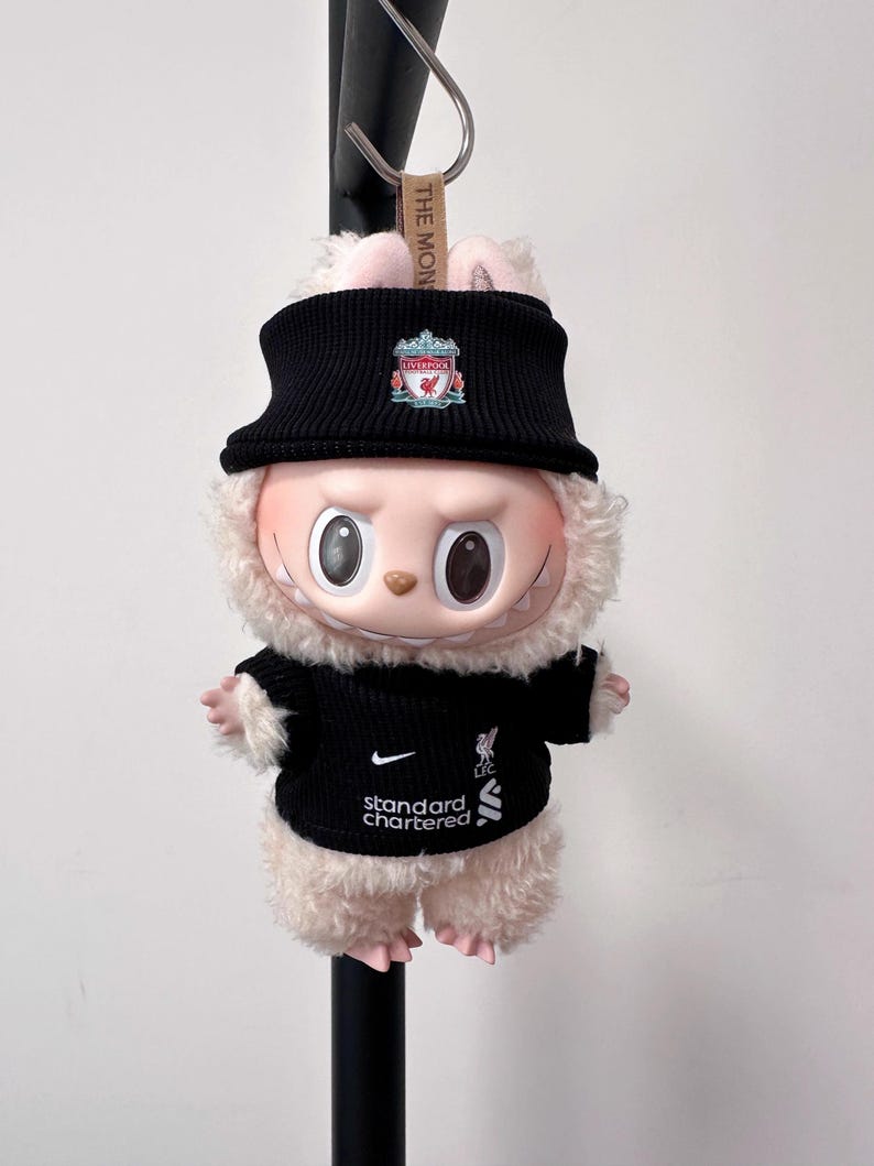 K&ouml;nnte beinhalten: Ein Stofftier-Hase mit schwarzem Strickhut und Pullover mit dem Logo des Liverpool Football Club. Der Pullover zeigt auch das Nike-Logo und den Text "Standard Chartered". Das Spielzeug hat ein hellrosa Gesicht und flauschiges Fell.