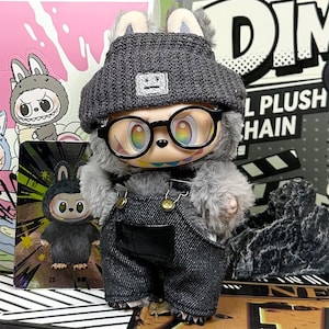 K&ouml;nnte beinhalten: Eine Pl&uuml;schtierfigur eines Kaninchens mit grauer M&uuml;tze, schwarz umrandeter Brille und Jeans-Latzhose. Die Figur hat regenbogenfarbene Augen und ein aufgesticktes L&auml;cheln. Der Hintergrund umfasst andere Spielzeugverpackungen und eine rosa-wei&szlig;e Grafik.