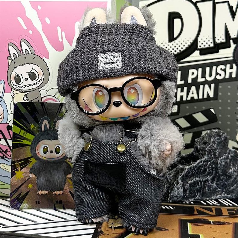 K&ouml;nnte beinhalten: Eine Pl&uuml;schtierfigur eines Kaninchens mit grauer M&uuml;tze, schwarz umrandeter Brille und Jeans-Latzhose. Die Figur hat regenbogenfarbene Augen und ein aufgesticktes L&auml;cheln. Der Hintergrund umfasst andere Spielzeugverpackungen und eine rosa-wei&szlig;e Grafik.